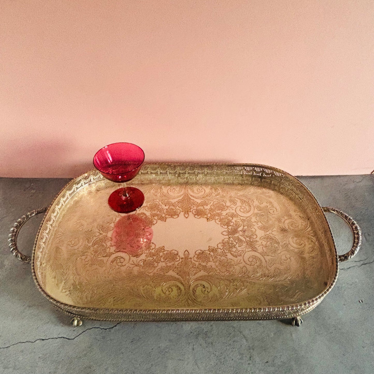 An ornate rectangular silver tray with decorative scrollwork and two side handles, shown on a gray surface against a pink wall. A small red glass bowl is placed on top of the tray.