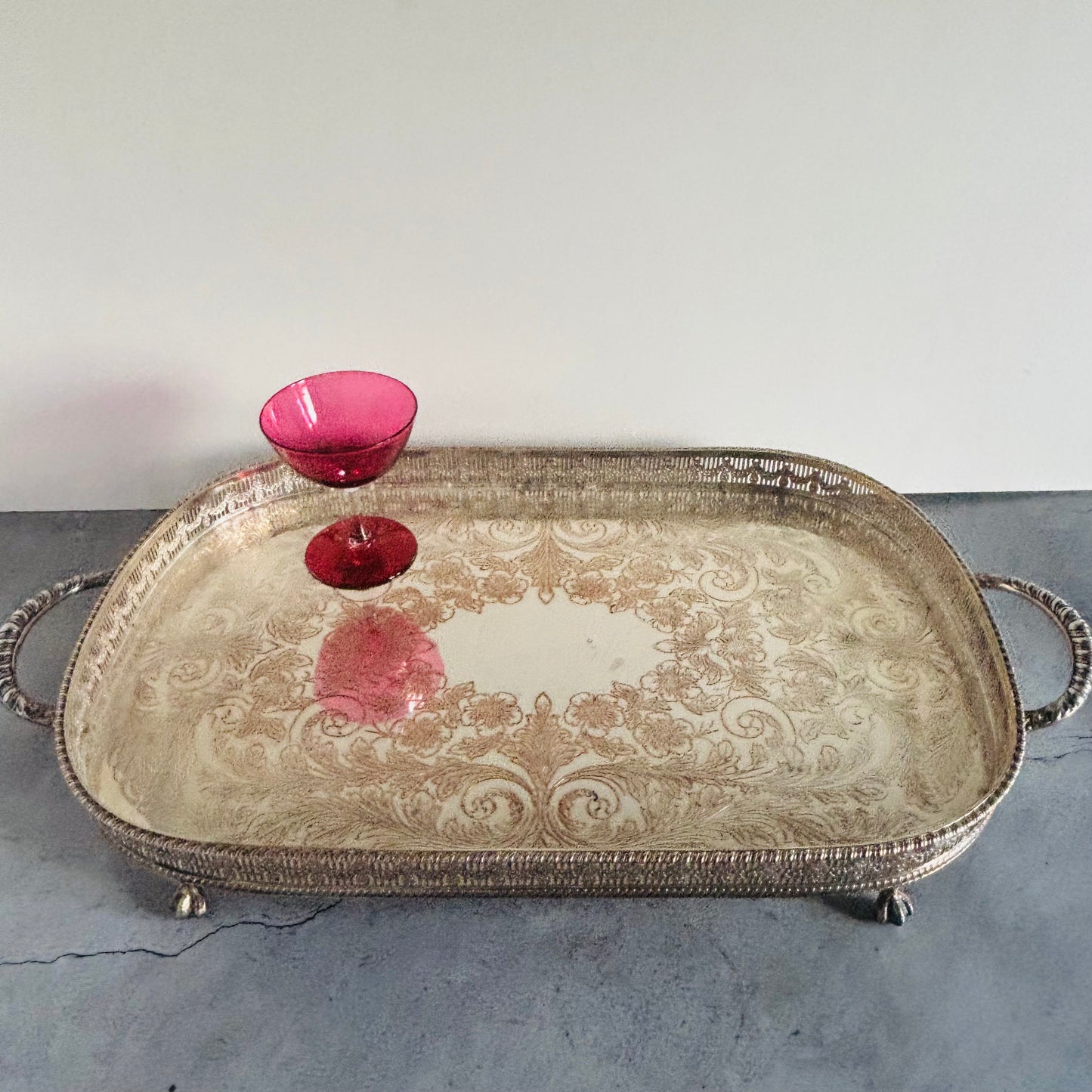 An ornate silver-toned metal serving tray with decorative scrollwork and loop handles, shown on a gray surface against a white wall. A small pink glass bowl is placed on the tray.