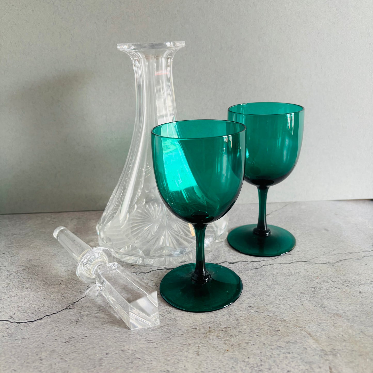 A set of vintage glassware including two emerald green stemmed wine glasses, a clear cut-crystal decanter, and a matching clear glass stopper, arranged on a light gray stone surface against a neutral gray background.