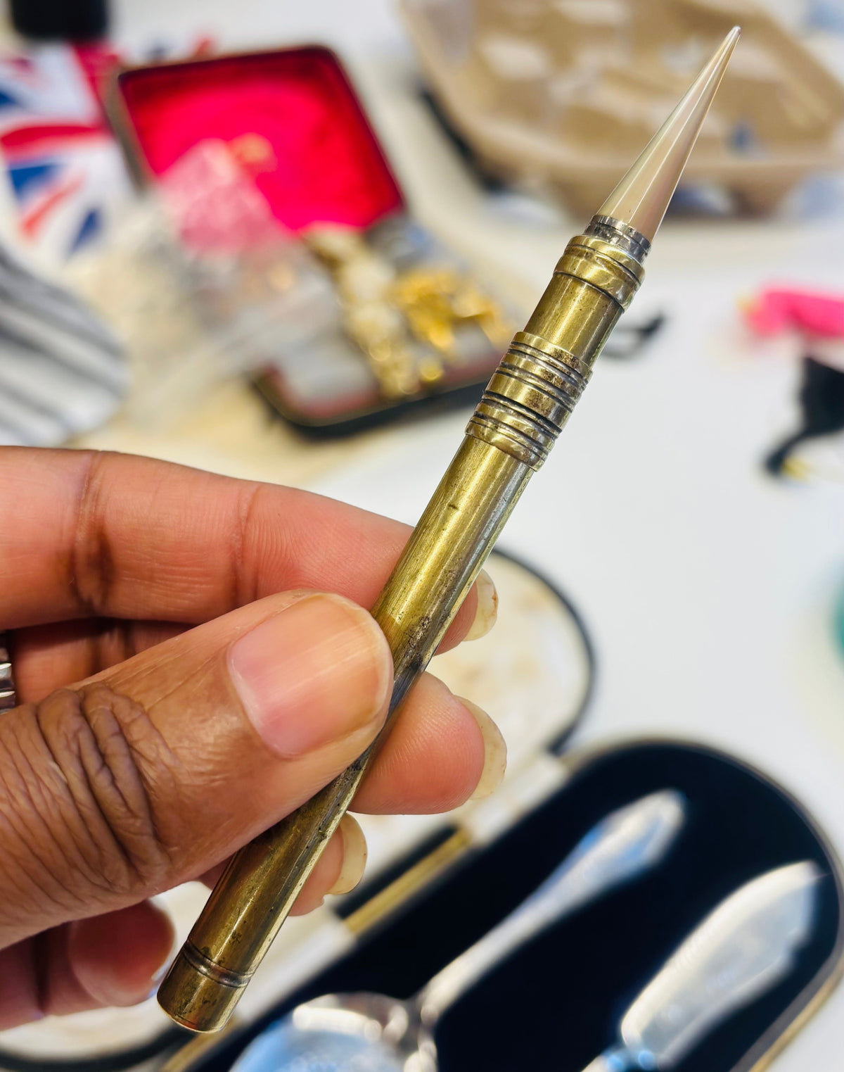 A hand holding an Antique Agate Gold Leaf Burnishing Tool with a pointed tip, with a silver spoon and knife in a black case blurred in the foreground.