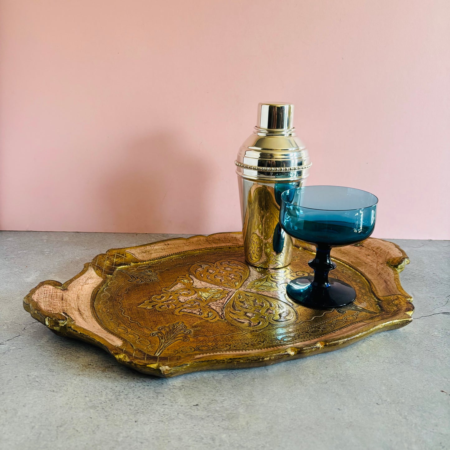Decorative vintage florentine tray with a silver cocktail shaker and blue glass on a pink background
