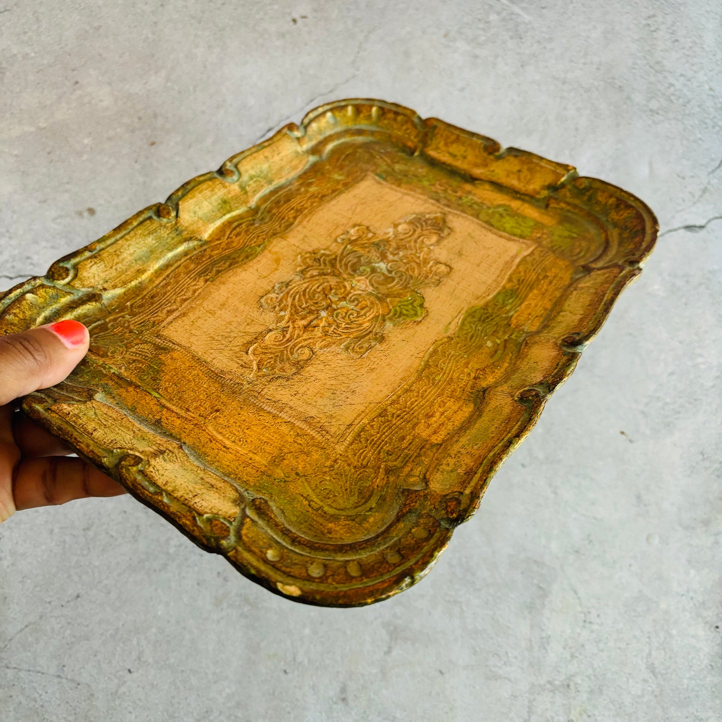 Decorative gold florentine tray with intricate designs held by a hand on a light gray surface