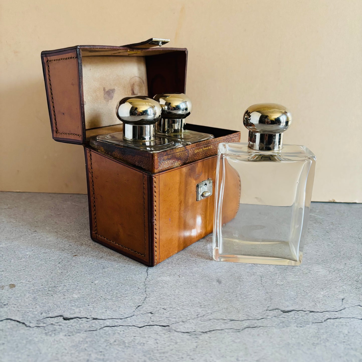 Vintage leather case with silver knobs and a clear glass bottle on a concrete floor.