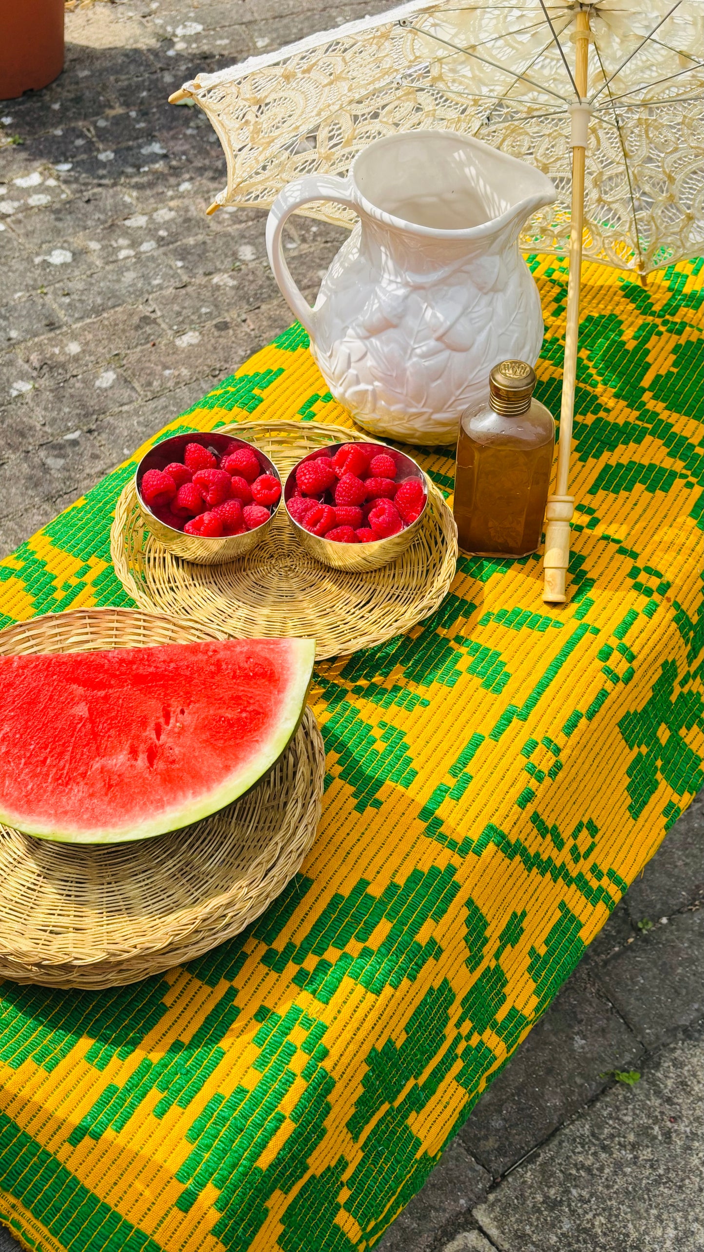 Table with a yellow and green patterned tablecloth, watermelon, raspberries, and a pitcher.