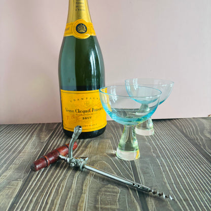 Bottle of Veuve Clicquot champagne with two cocktail glasses and a corkscrew on a wooden surface.