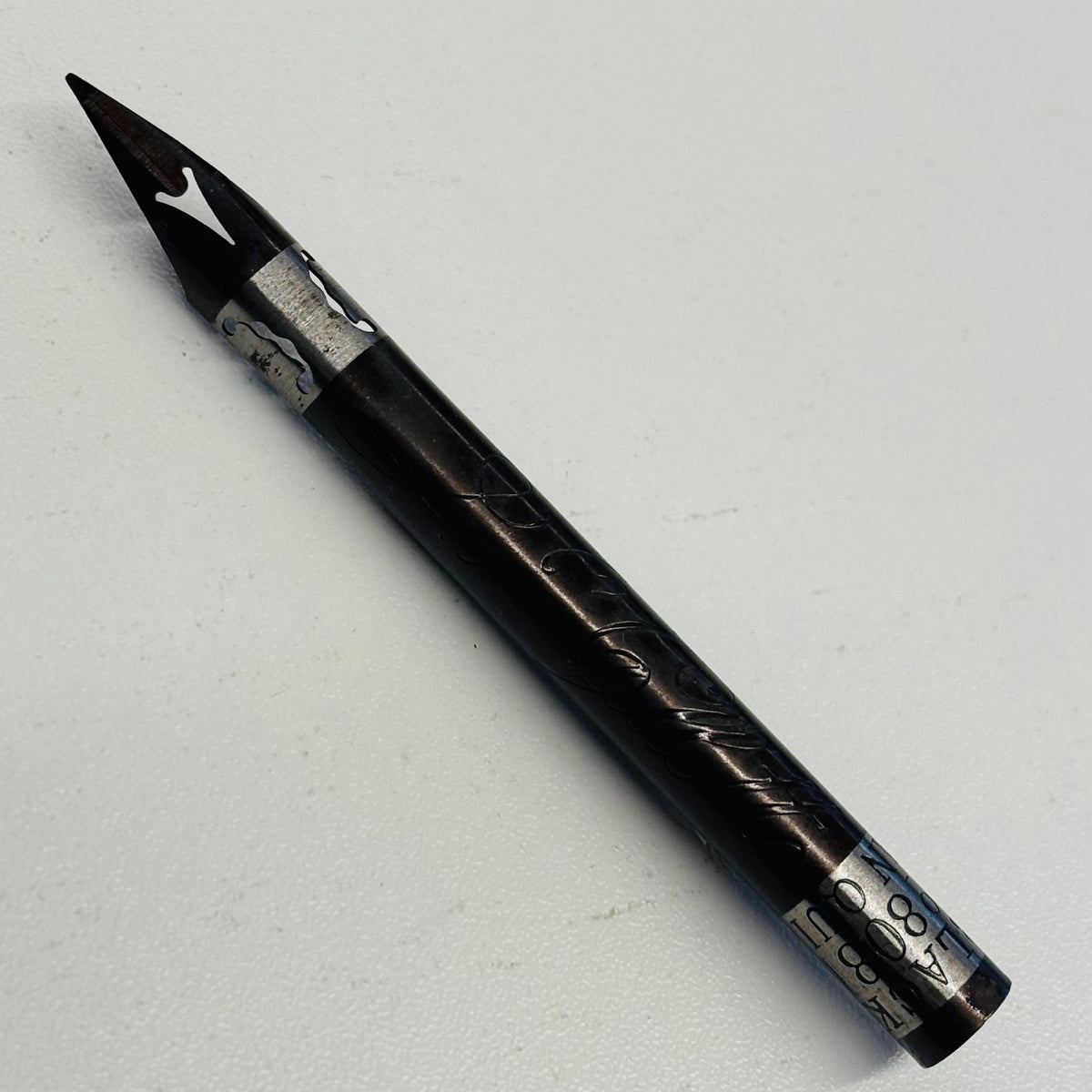 A black pen nib with silver engraved details and a silver nib, photographed diagonally on a light gray background.