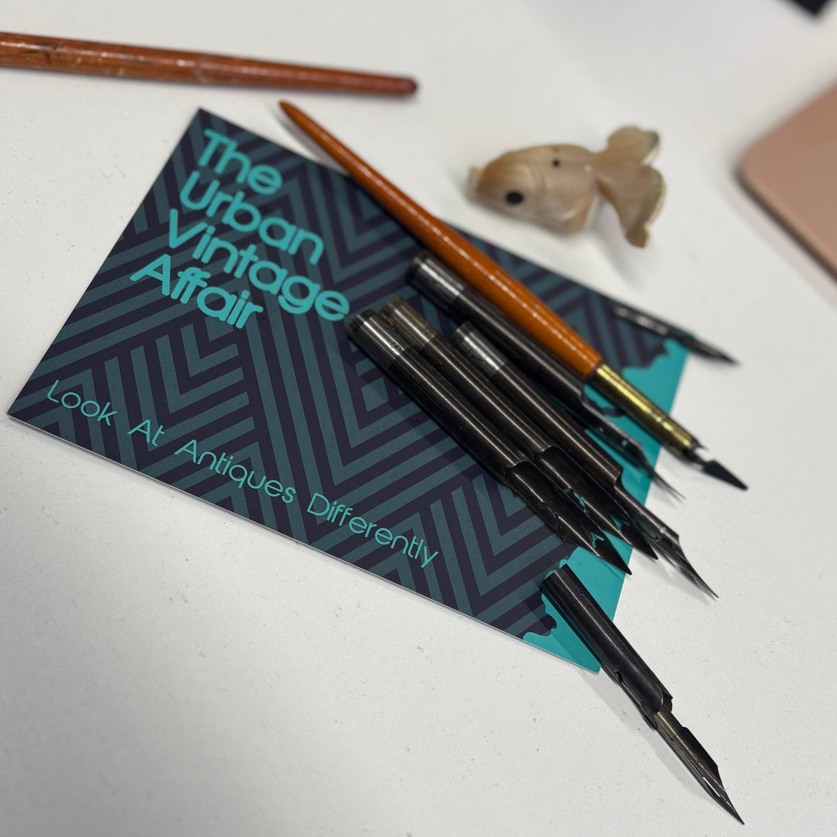 A set of vintage-style calligraphy or paint brushes with orange and black handles, displayed on a dark patterned card that reads 'The Urban Vintage Affair' on a white desk.