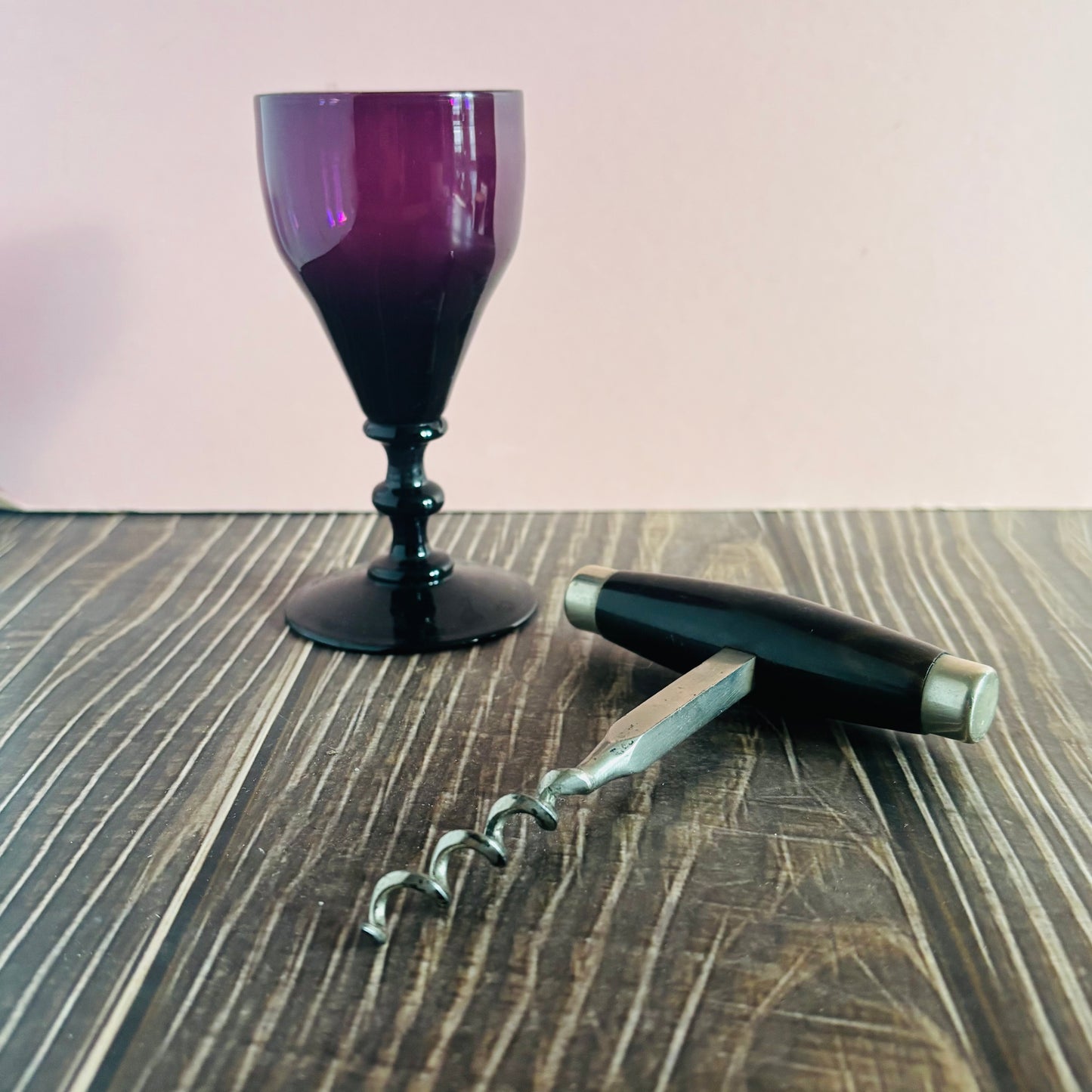 Purple wine glass, corkscrew, and black pen on a wooden surface with a pink background