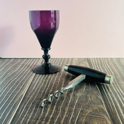 Purple wine glass, corkscrew, and black pen on a wooden surface with a pink background