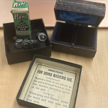A vintage Feraei black ink bottle set displayed on a desk with an open presentation box showing the bottle, ink stones, and an instruction card, with a blurred laptop in the background.