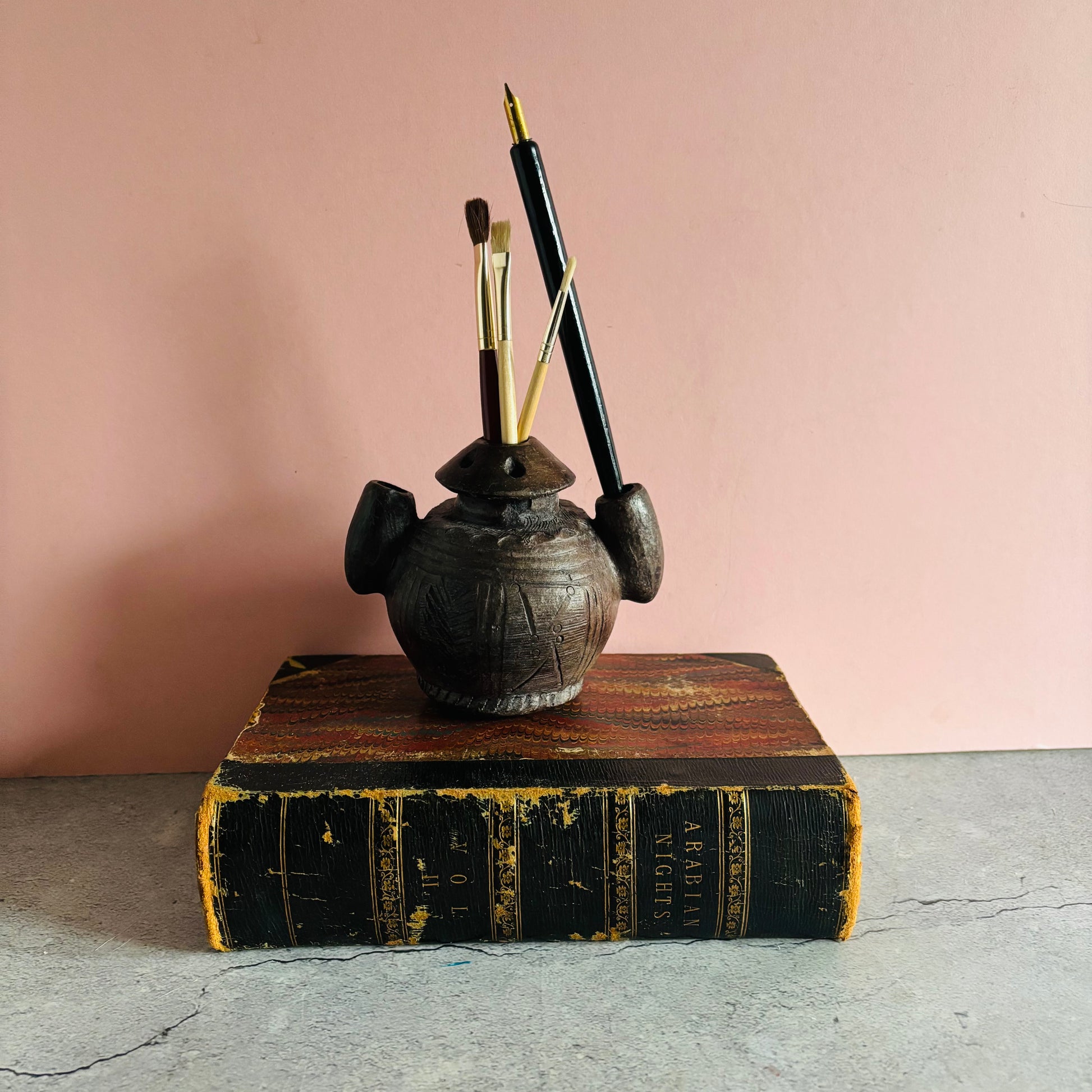 Antique African Clay Ink Pot From Kano Nigeria with pens on an old book against a pink wall.