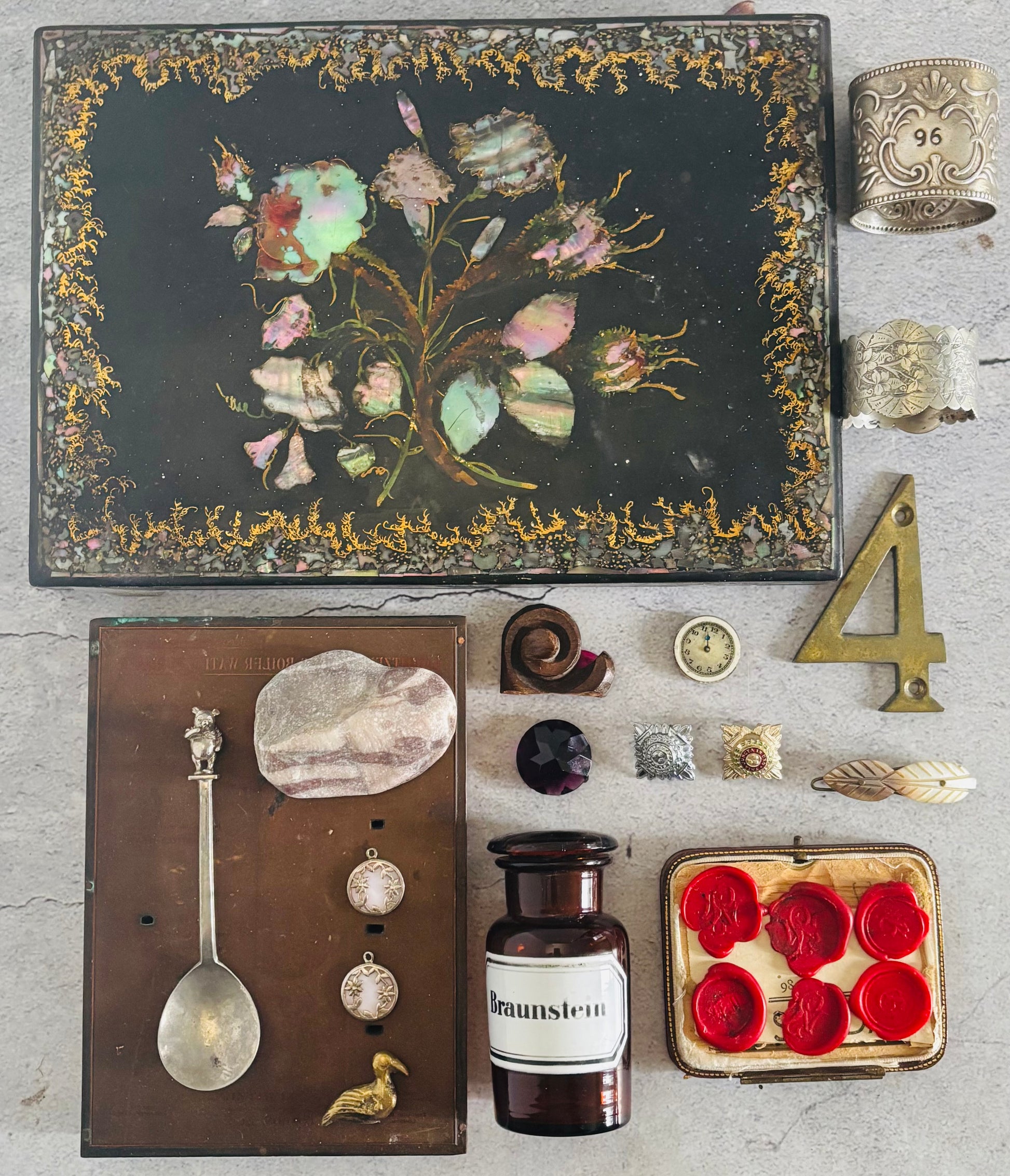 Collection of vintage items including a decorative box with floral design, a spoon, a bottle, and other small objects on a textured surface.