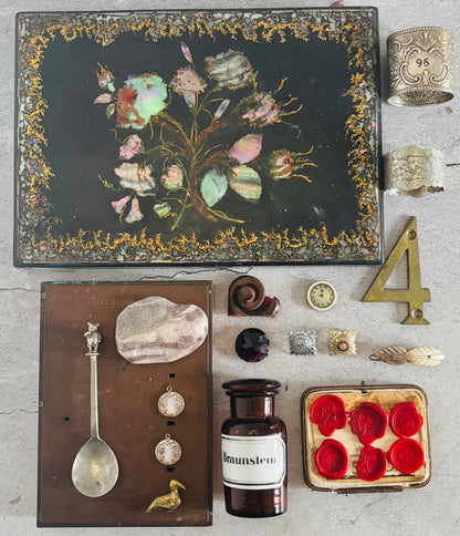 Collection of vintage items including a decorative box with floral design, a spoon, a bottle, and other small objects on a textured surface.