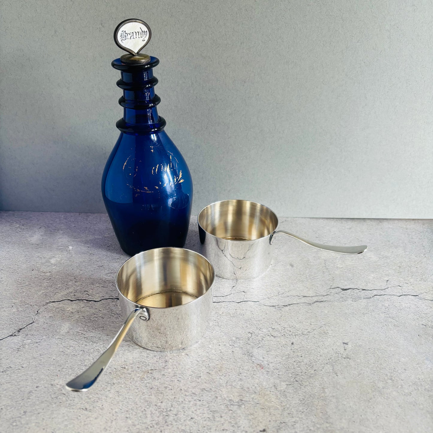 Blue bandy glass decanter with a silver lid and two silver brandy pans on a textured surface.