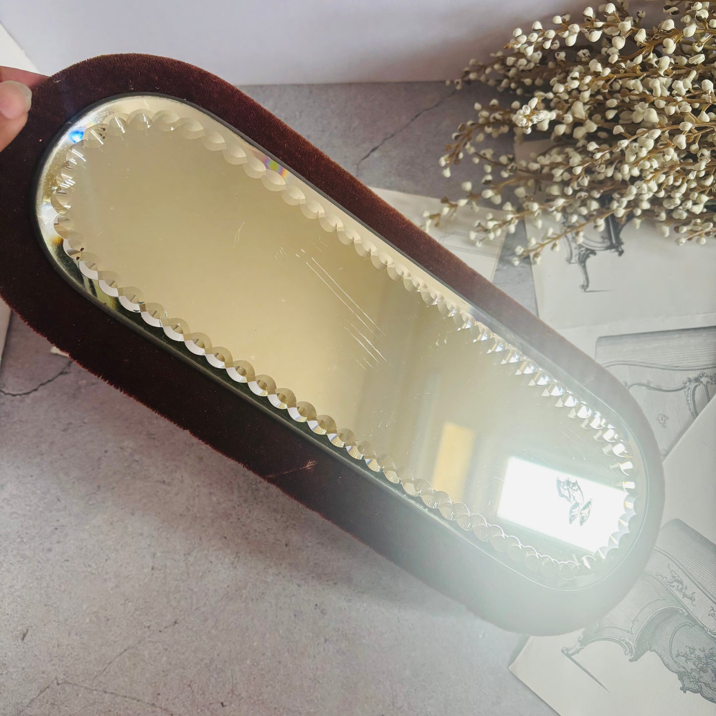 Small oval mirror with decorative frame held by a hand on a light-colored surface.