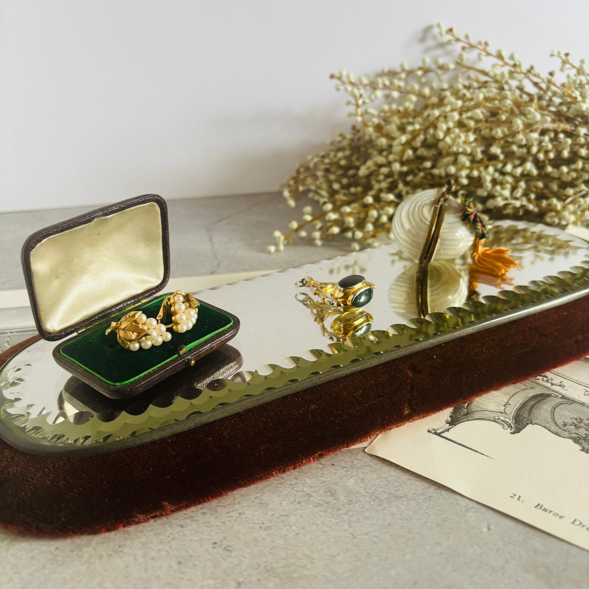 Gold jewelry set on a reflective surface with a vintage book and dried flowers in the background.