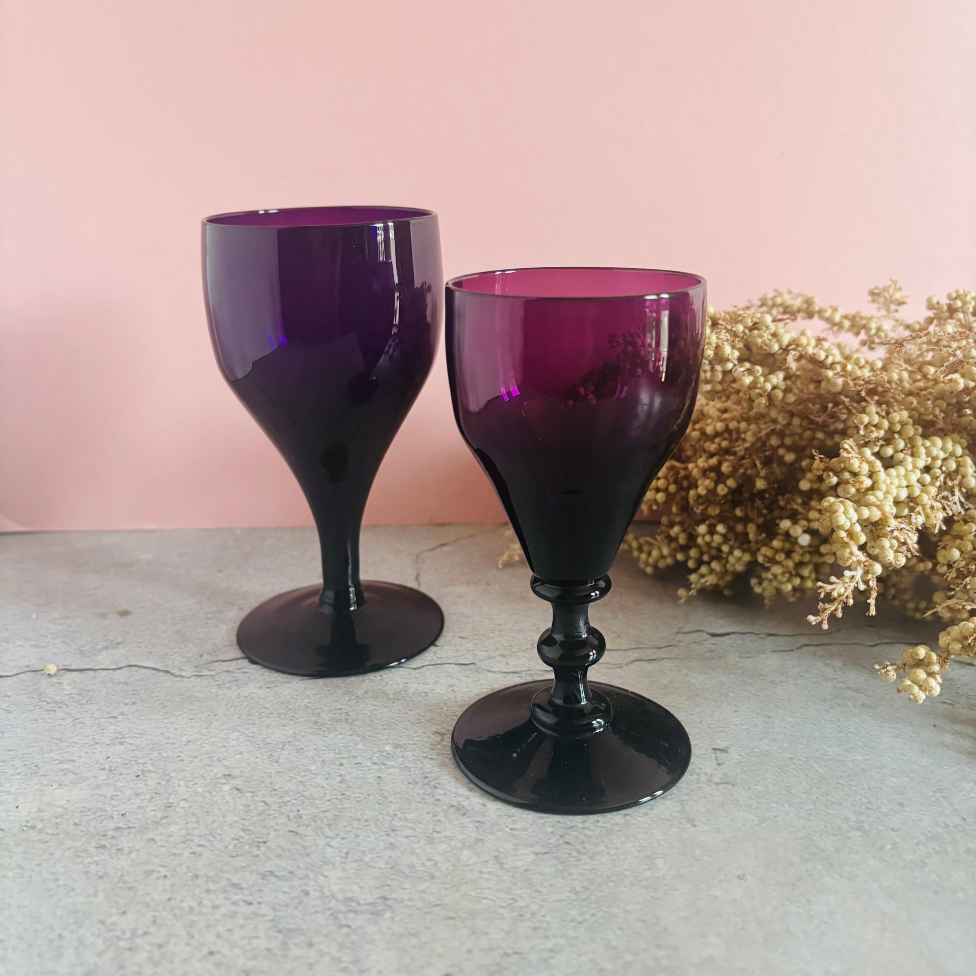 Two purple wine glasses on a light gray surface with a pink background and dried flowers.