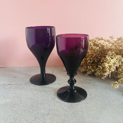 Two purple wine glasses on a light gray surface with a pink background and dried flowers.