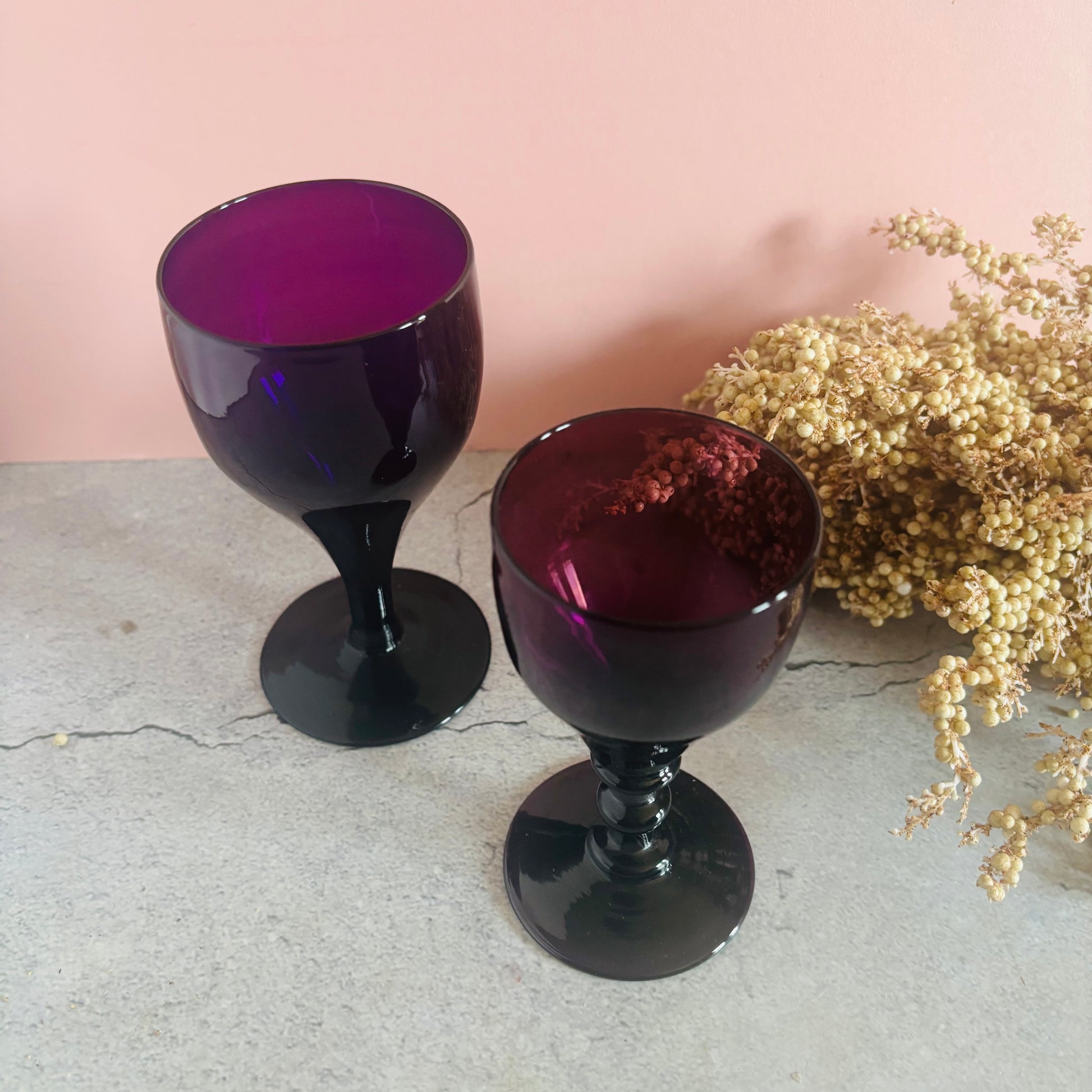 Two purple glass goblets on a light surface with a pink background and dried flowers.