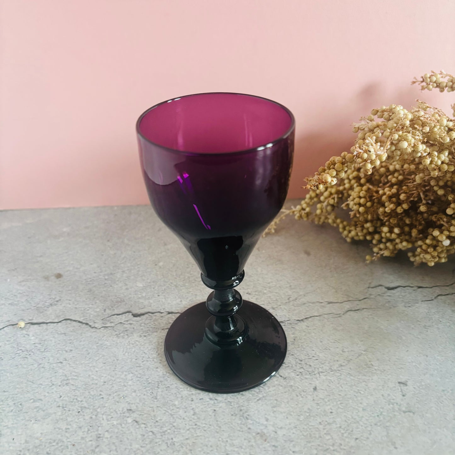 Amethyst-colored goblet on a concrete surface with a pink wall and dried flowers in the background