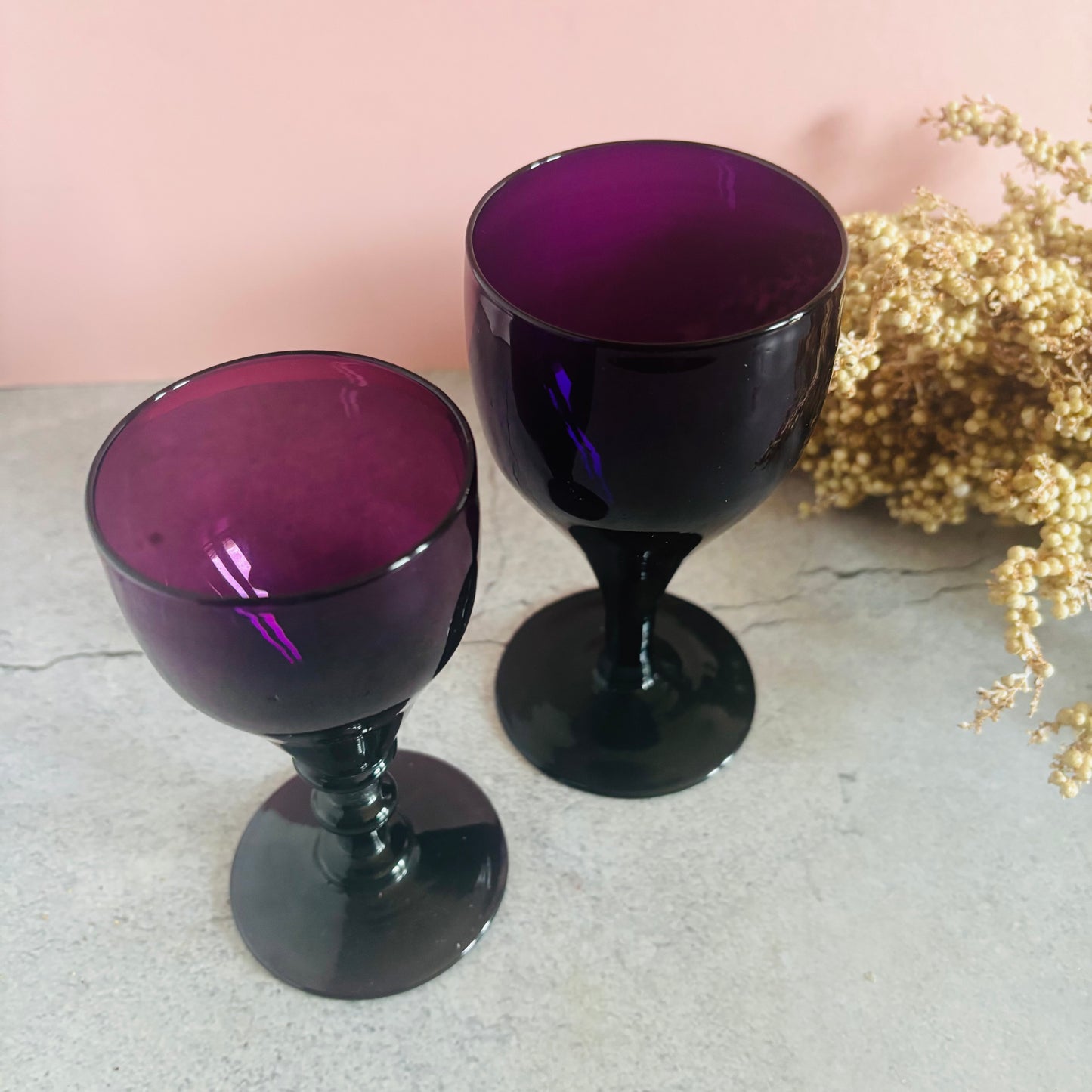 Two purple glass goblets on a light surface with a pink background and gold decorative elements.