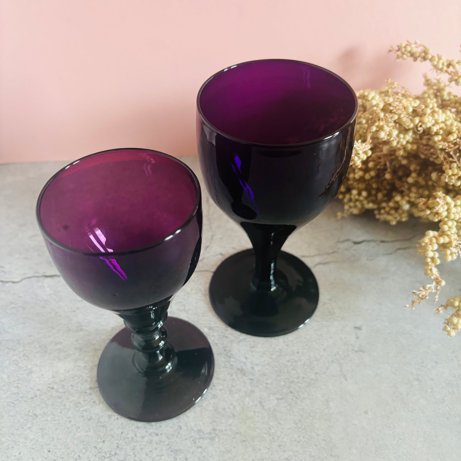 Two purple glass goblets on a light surface with a pink background and gold decorative elements.