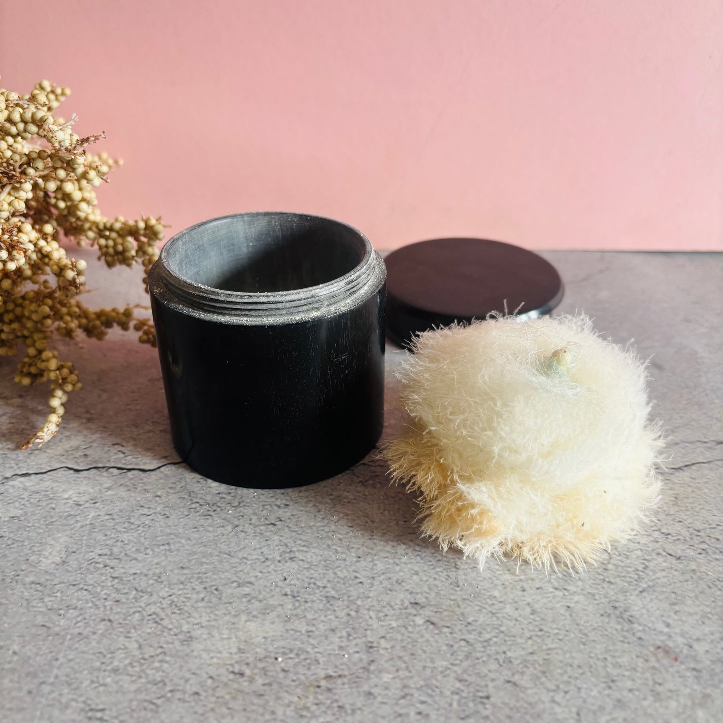 Black ebony container with a white fluffy puffer on a gray surface against a pink background