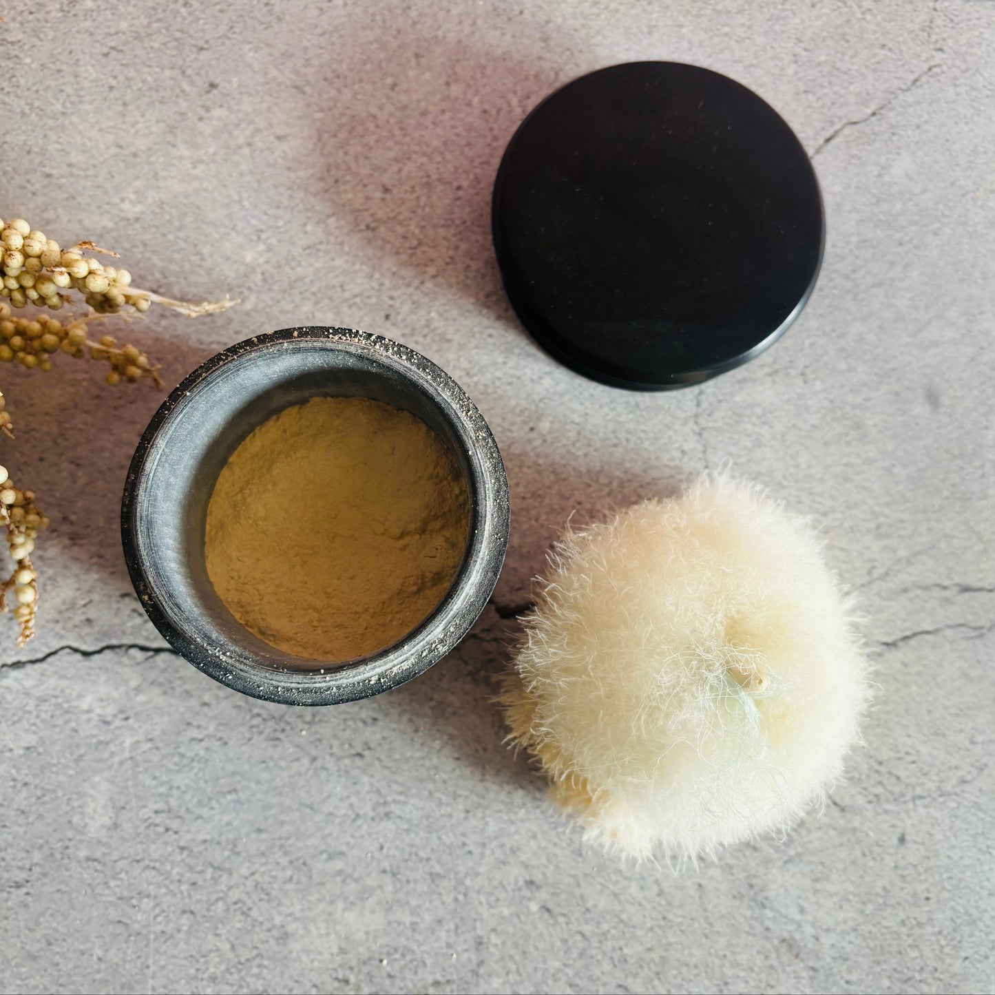 Open ebony container with  powder and black lid on a grey surface with dried plants.