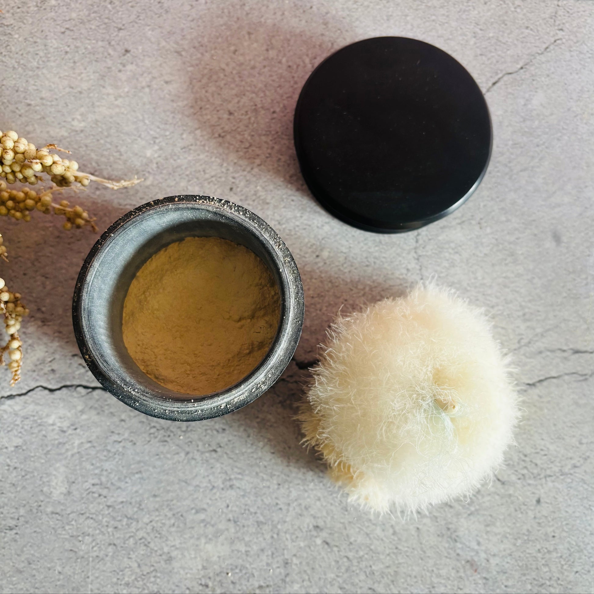 Open ebony container with  powder and black lid on a grey surface with dried plants.