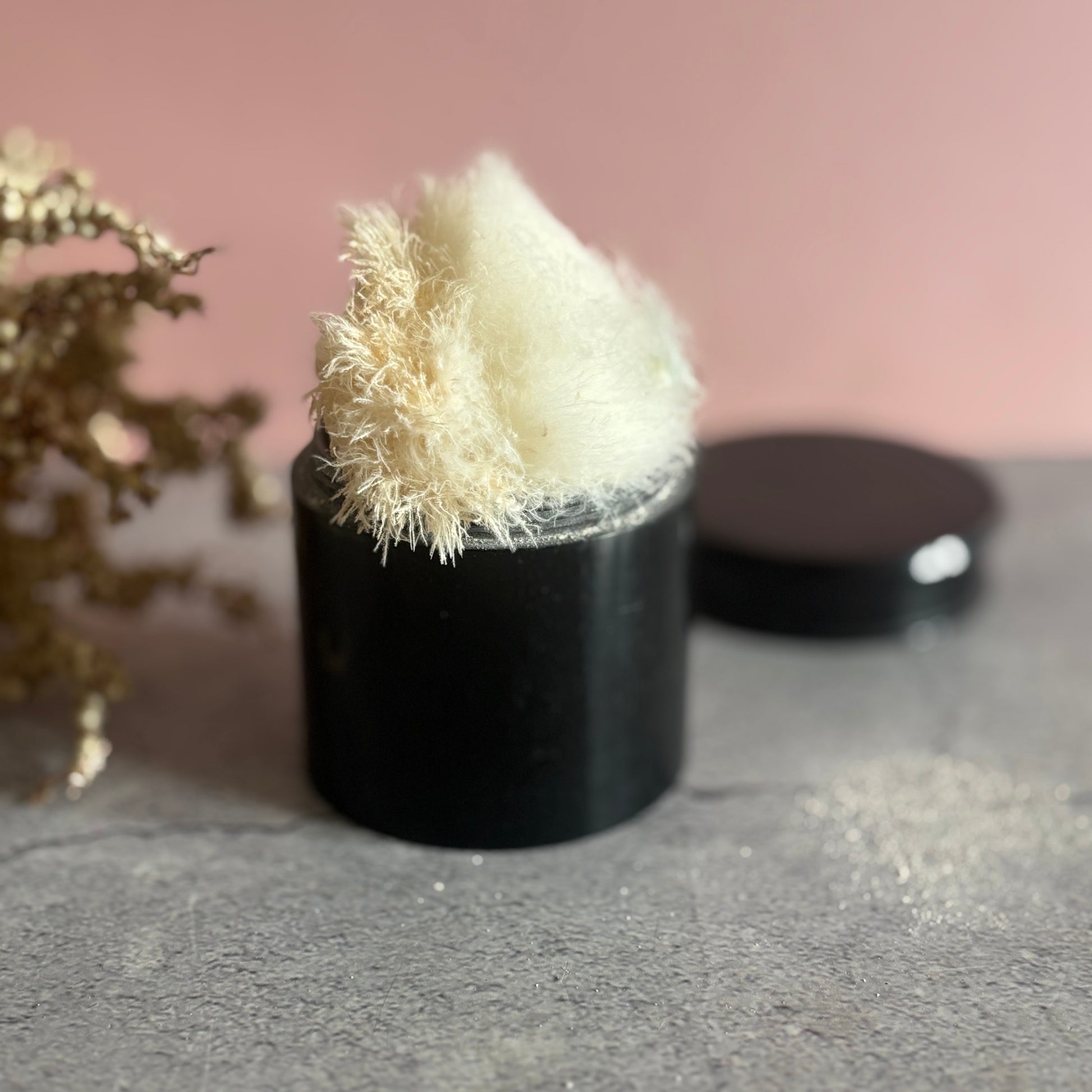 Black ebony container with a white fluffy puffer on a gray surface with a pink background