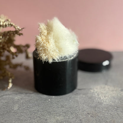 Black ebony container with a white fluffy puffer on a gray surface with a pink background