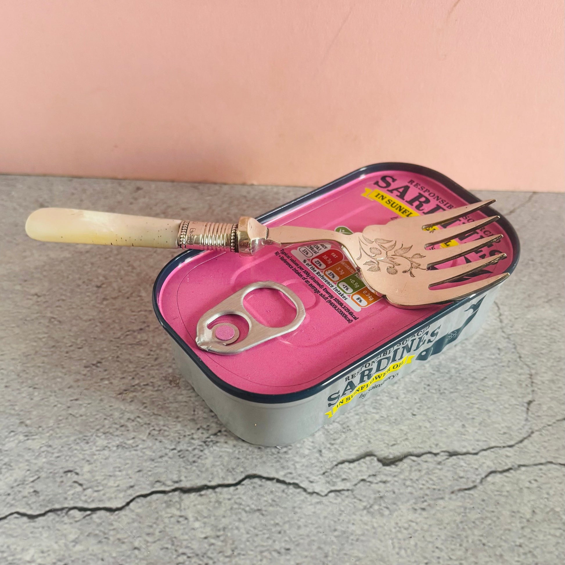 Sardine can with a fork and knife on a cracked surface with a pink background