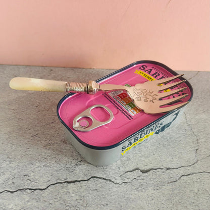 Sardine can with a fork and knife on a cracked surface with a pink background