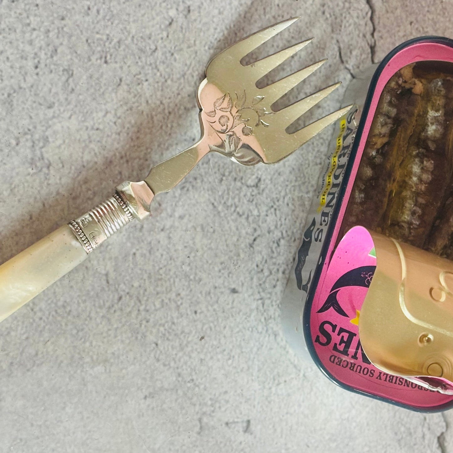 Antique Silver Sardine Fork With Mother Of Pearl Handle next to a can of sardines on a concrete surface