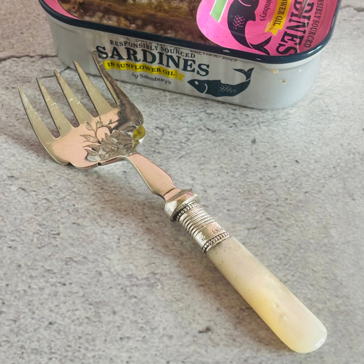 Antique Sardine fork with decorative handle next to a can of sardines on a concrete surface