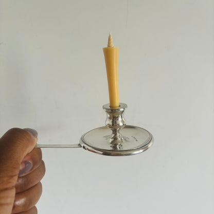 Hand holding a silver candle holder with a lit yellow candle against a plain background