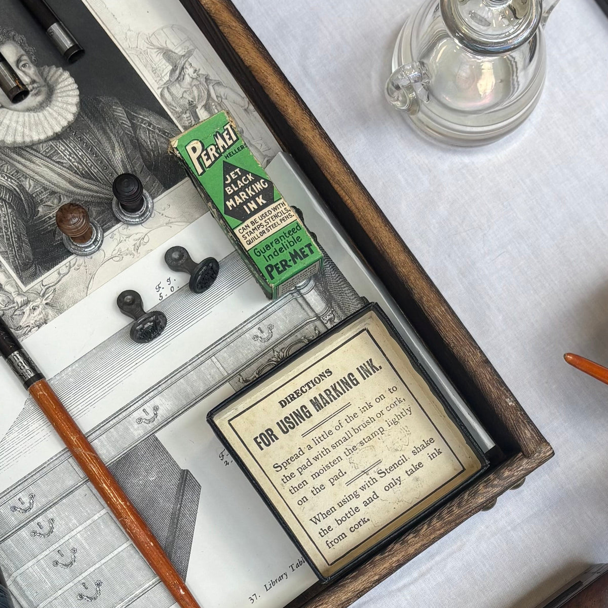 An overhead flat-lay of vintage stationery and writing tools arranged on a white surface, including a wooden ink tray with a quill, small glass bottles, a decorative box, an open book, and a leather-bound notebook.