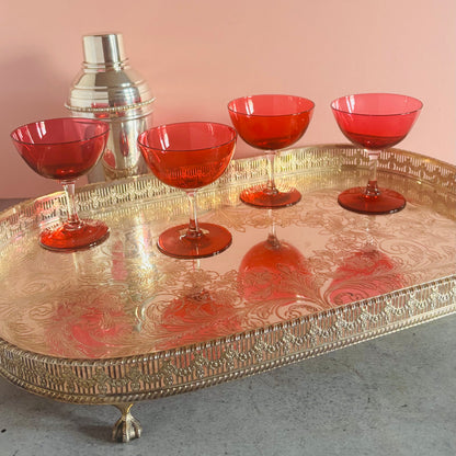 A decorative silver-toned tray with engraved detailing holds four translucent red cocktail glasses and a silver shaker against a solid pink wall.