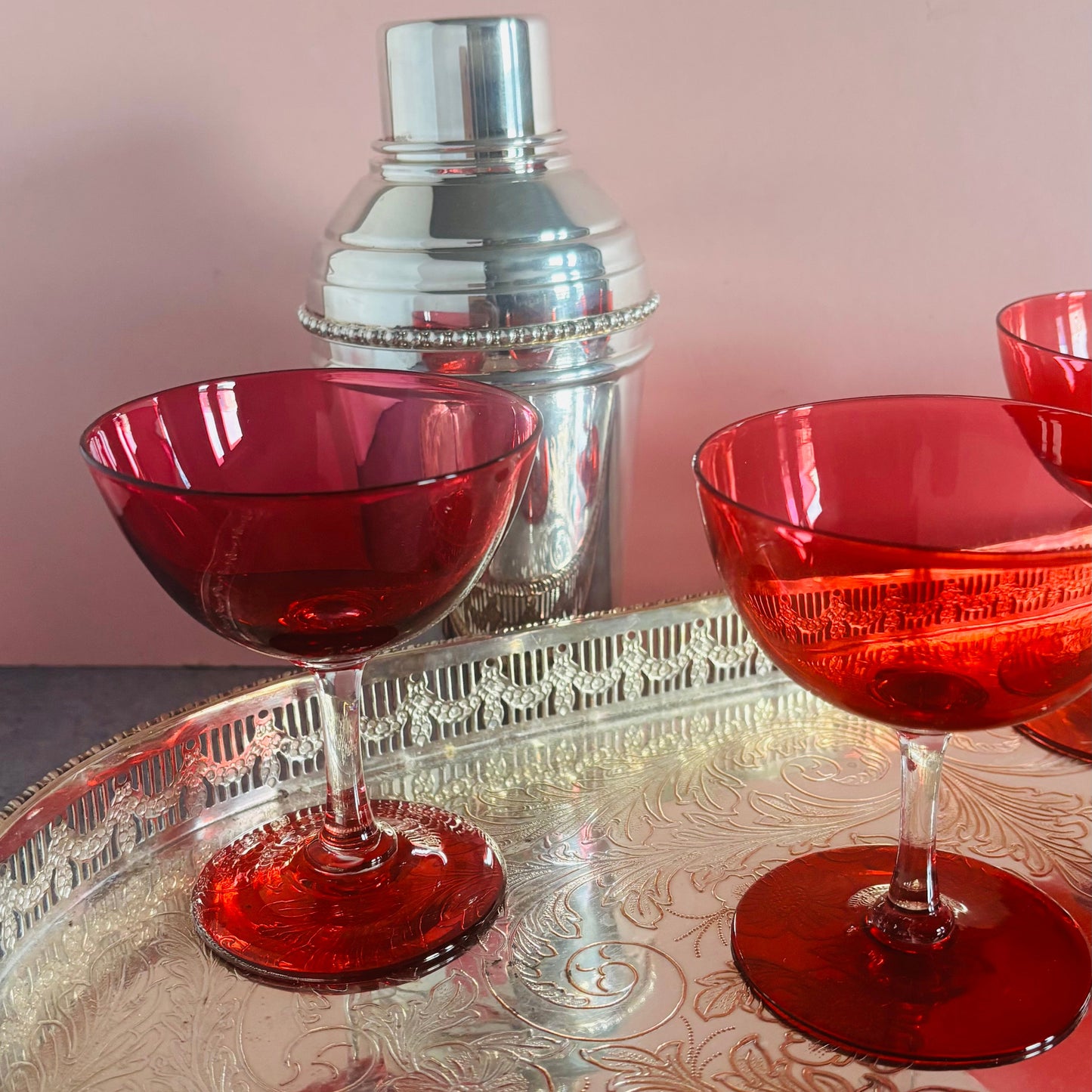 A antique cocktail shaker with a silver metal body and lid, positioned behind two cranberry glass coupe glasses on a patterned silver tray against a solid pink background.