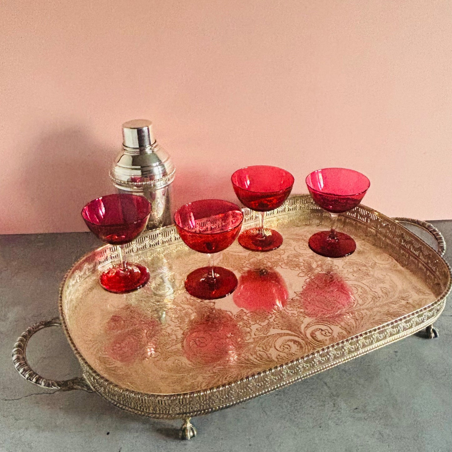 A decorative oval silver tray with ornate handles and feet, holding six red glass cocktail glasses and a small silver shaker, set against a pink wall and gray surface.