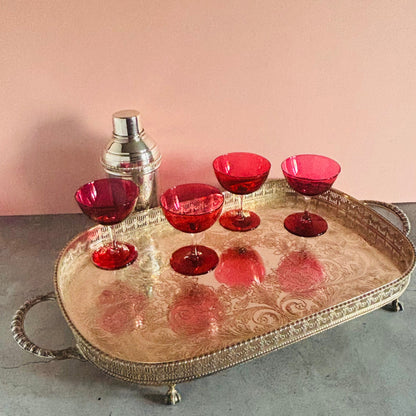 A decorative oval silver tray with ornate handles and feet, holding six red glass cocktail glasses and a small silver shaker, set against a pink wall and gray surface.
