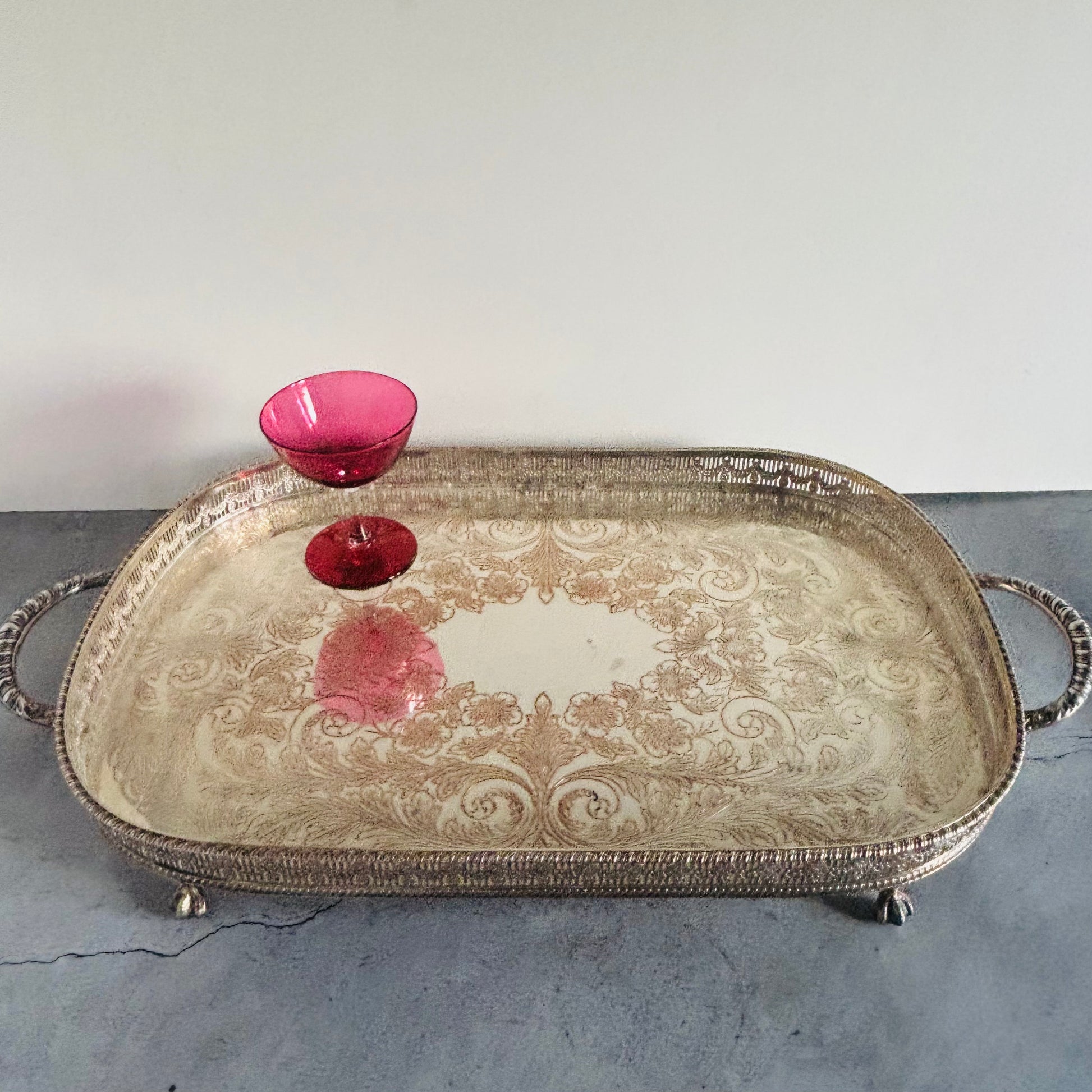 An ornate silver-toned metal serving tray with decorative scrollwork and loop handles, shown on a gray surface against a white wall. A small pink glass bowl is placed on the tray.