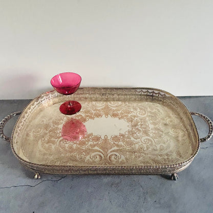 An ornate silver-toned metal serving tray with decorative scrollwork and loop handles, shown on a gray surface against a white wall. A small pink glass bowl is placed on the tray.