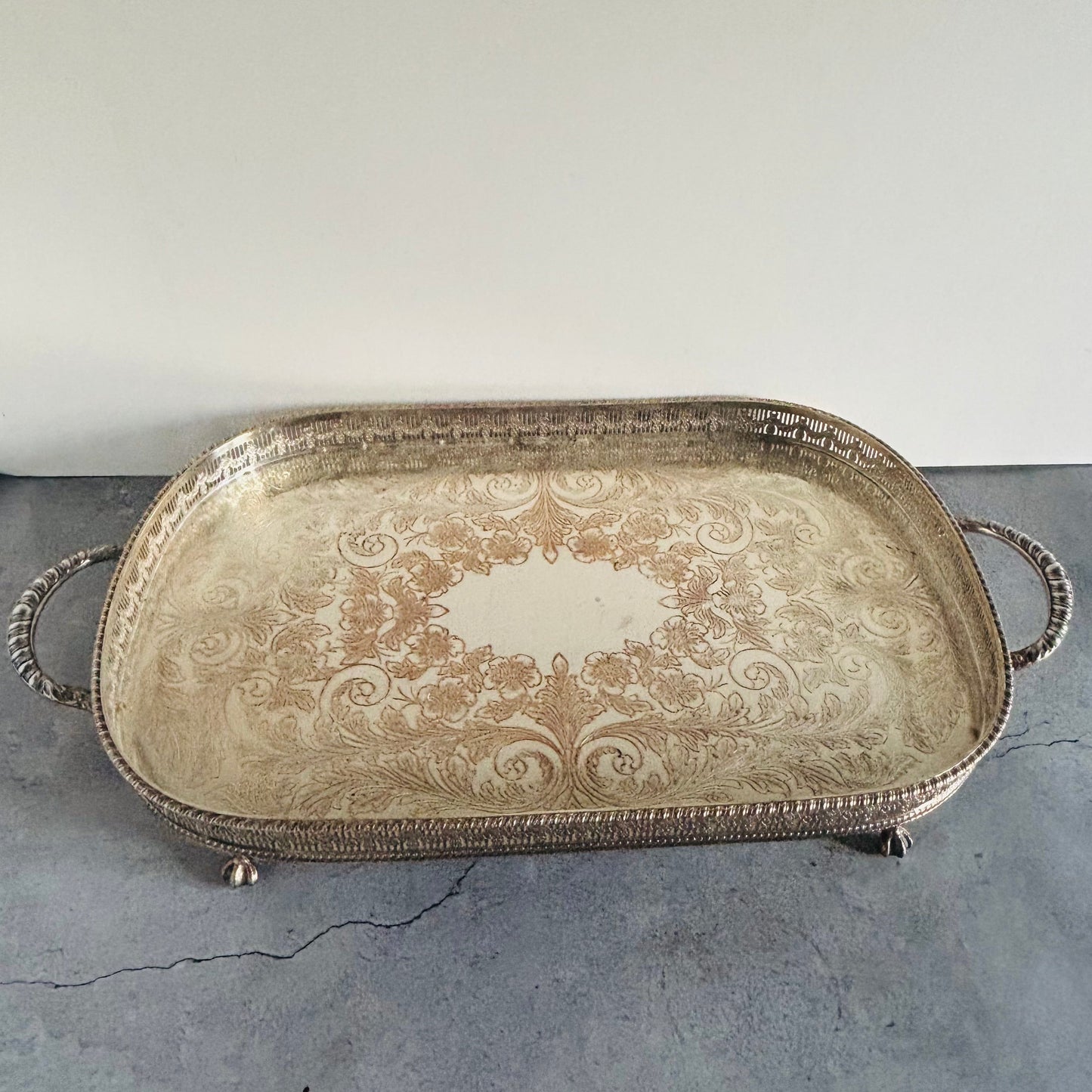 An ornate rectangular silver serving tray with decorative handles and feet, featuring an embossed damask pattern, resting on a gray concrete floor against a plain white wall.