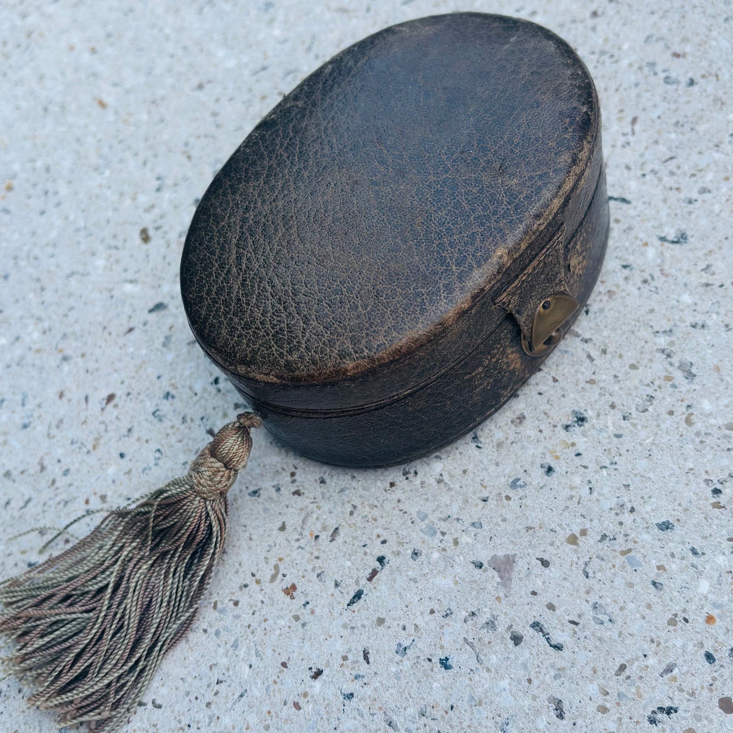 A small oval black leather case with a tassel, photographed on a speckled light gray concrete surface.
