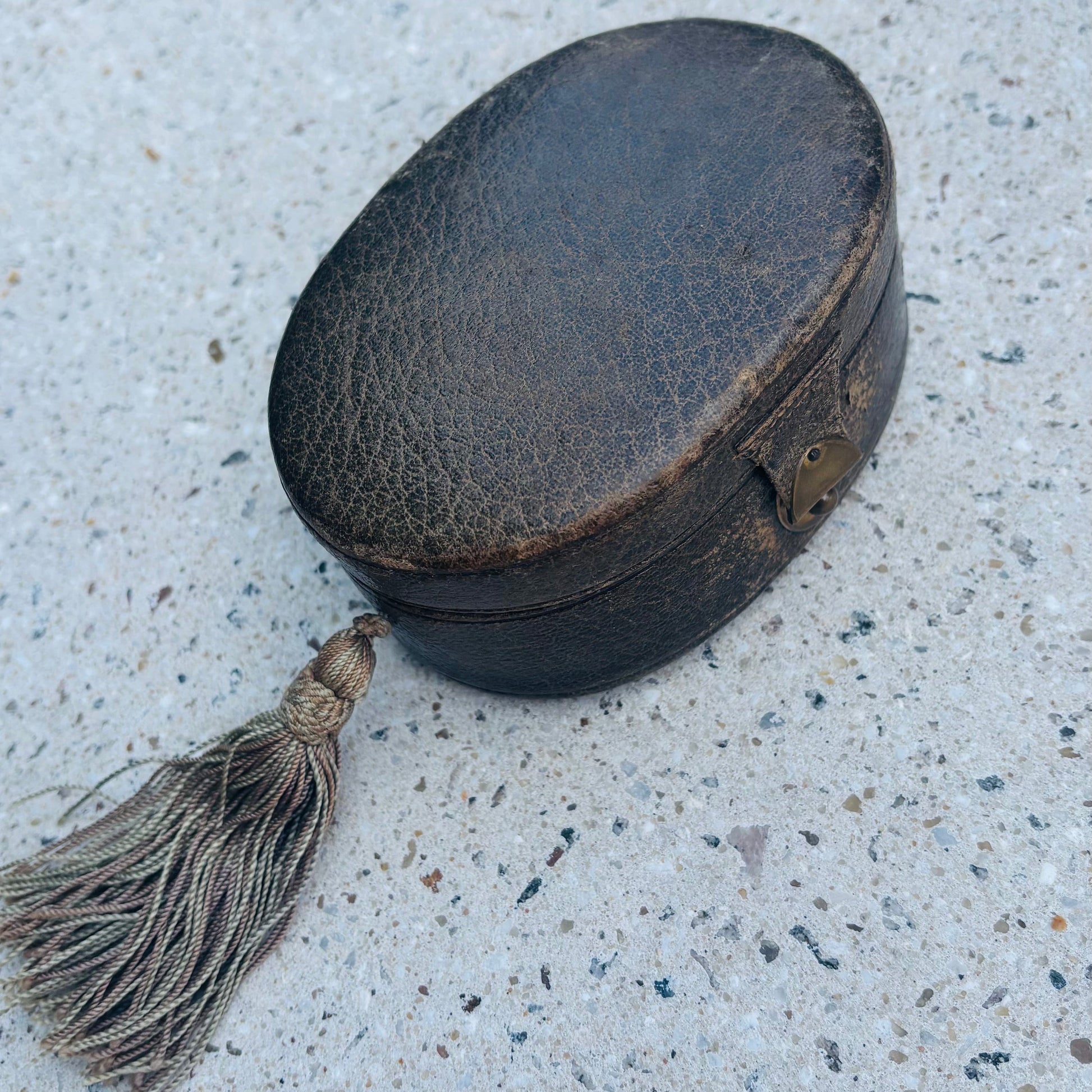 A small oval black leather case with a tassel, photographed on a speckled light gray concrete surface.