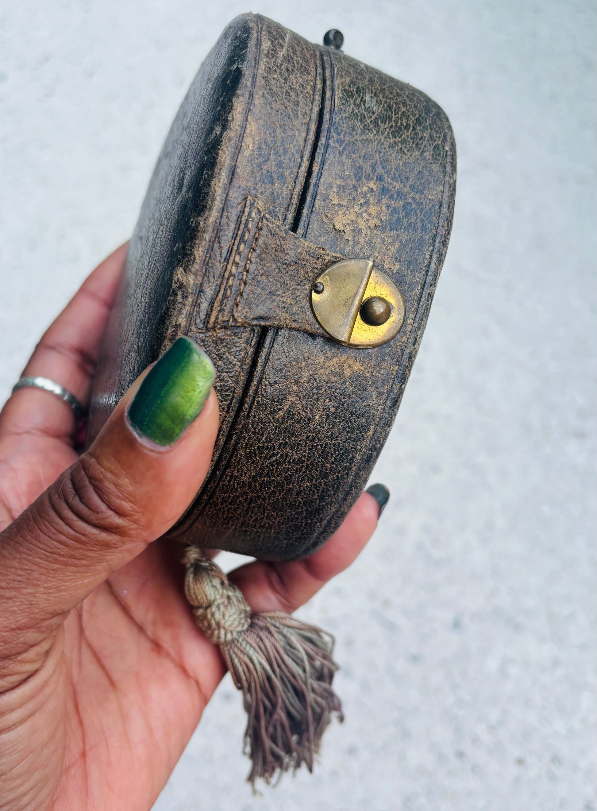 A hand with green nail polish holds a small, round, vintage-style dark brown leather pouch or case with a brass clasp and a tassel.