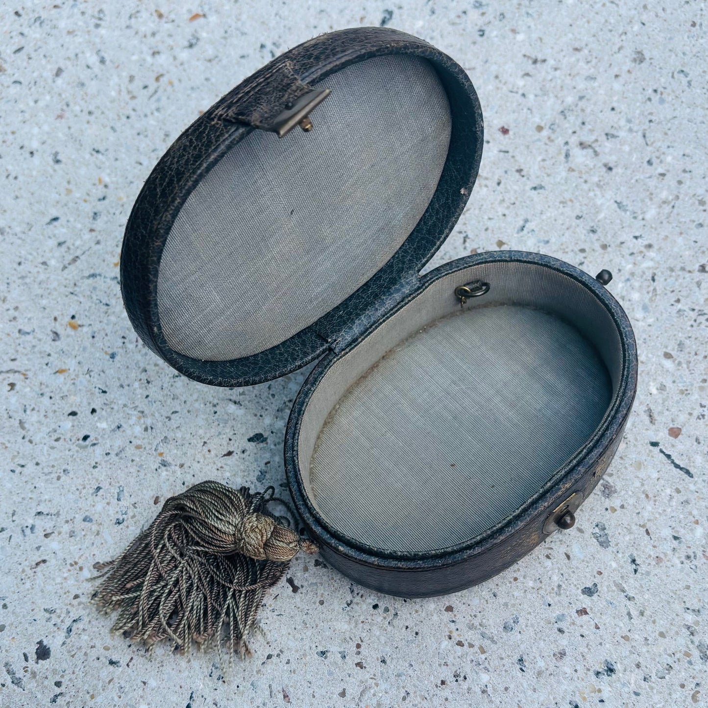 An antique oval leather case with a tassel, shown open on a speckled concrete surface.