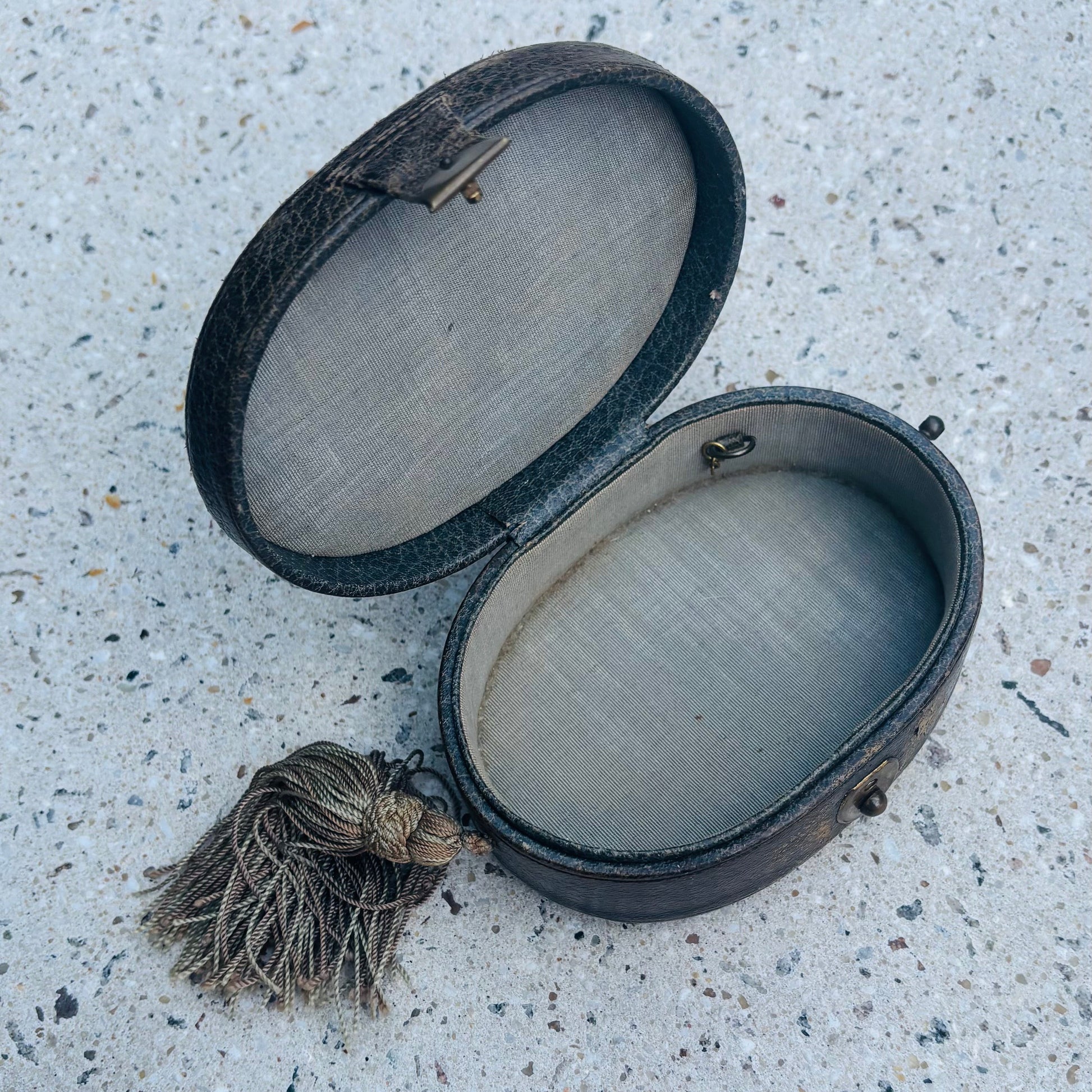 An antique oval leather case with a tassel, shown open on a speckled concrete surface.