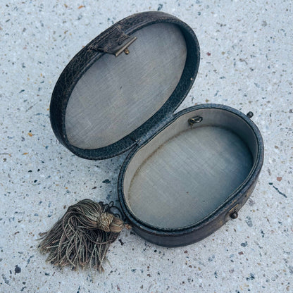 An antique oval leather case with a tassel, shown open on a speckled concrete surface.